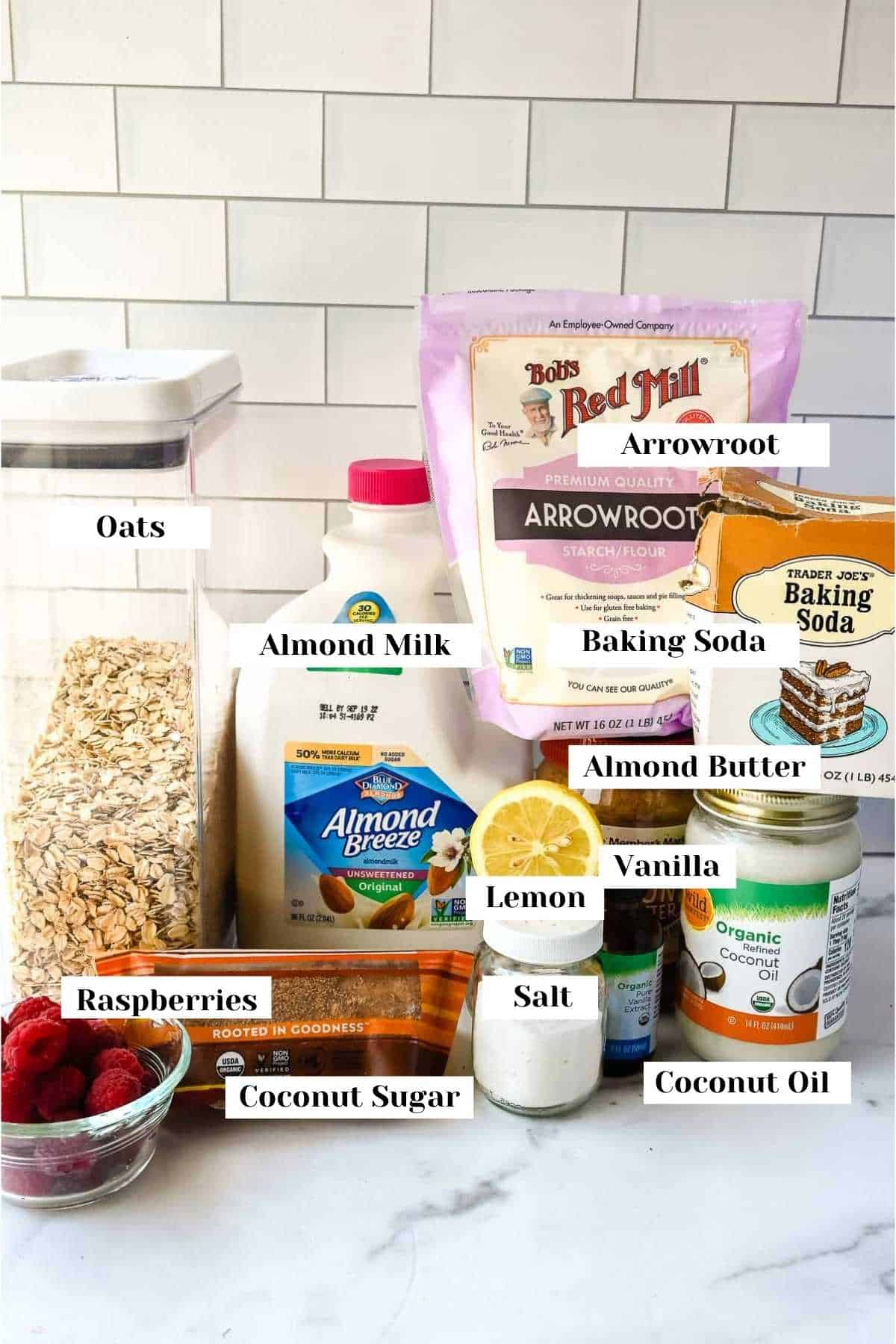 labeled ingredient photo showing oats, almond milk, raspberries, coconut sugar, salt, coconut oil, vanilla, lemon, almond butter, baking soda and arrowroot.