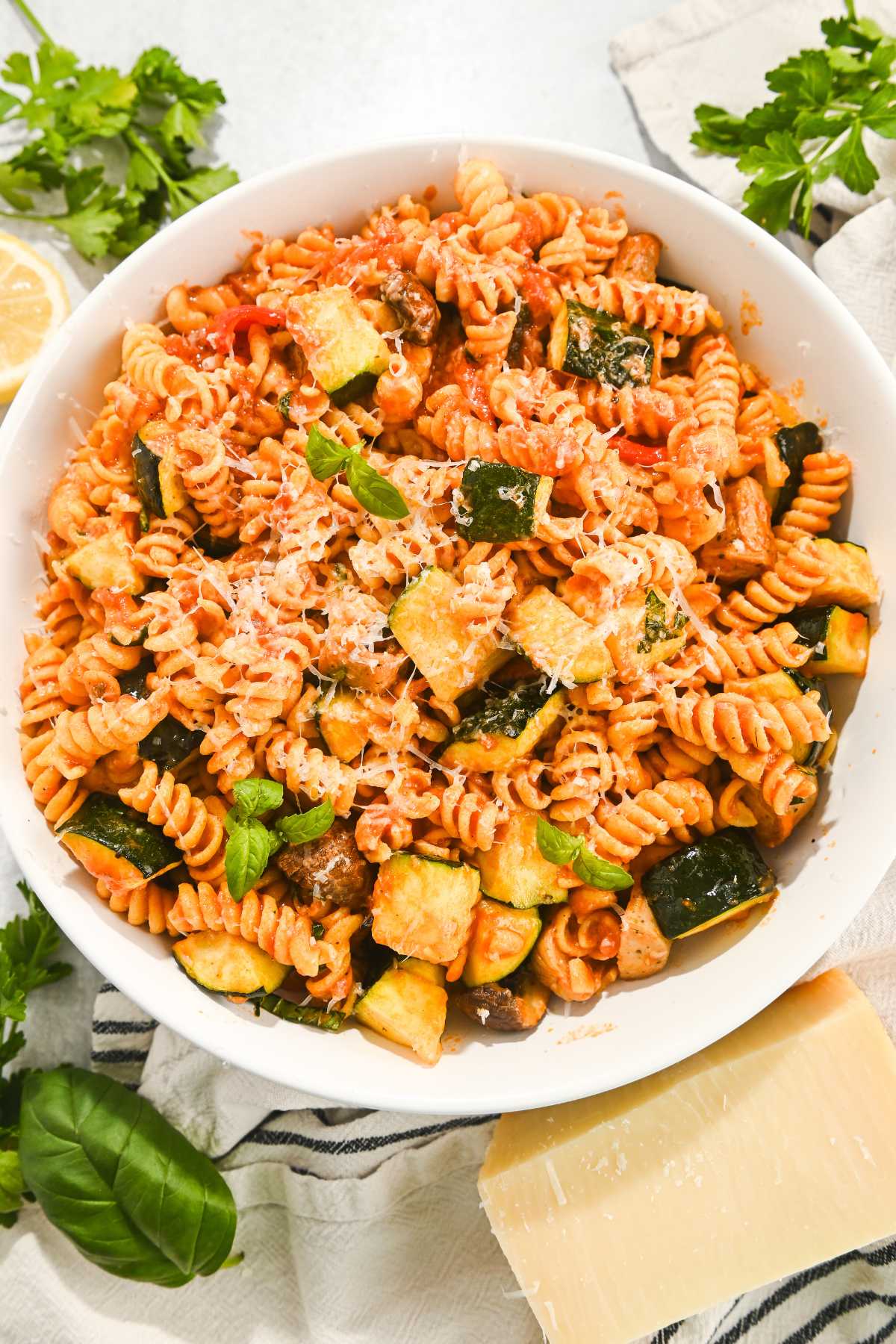 overhead view of chicken sausage pasta in a white bowl next to fresh herbs and Parmesan cheese.
