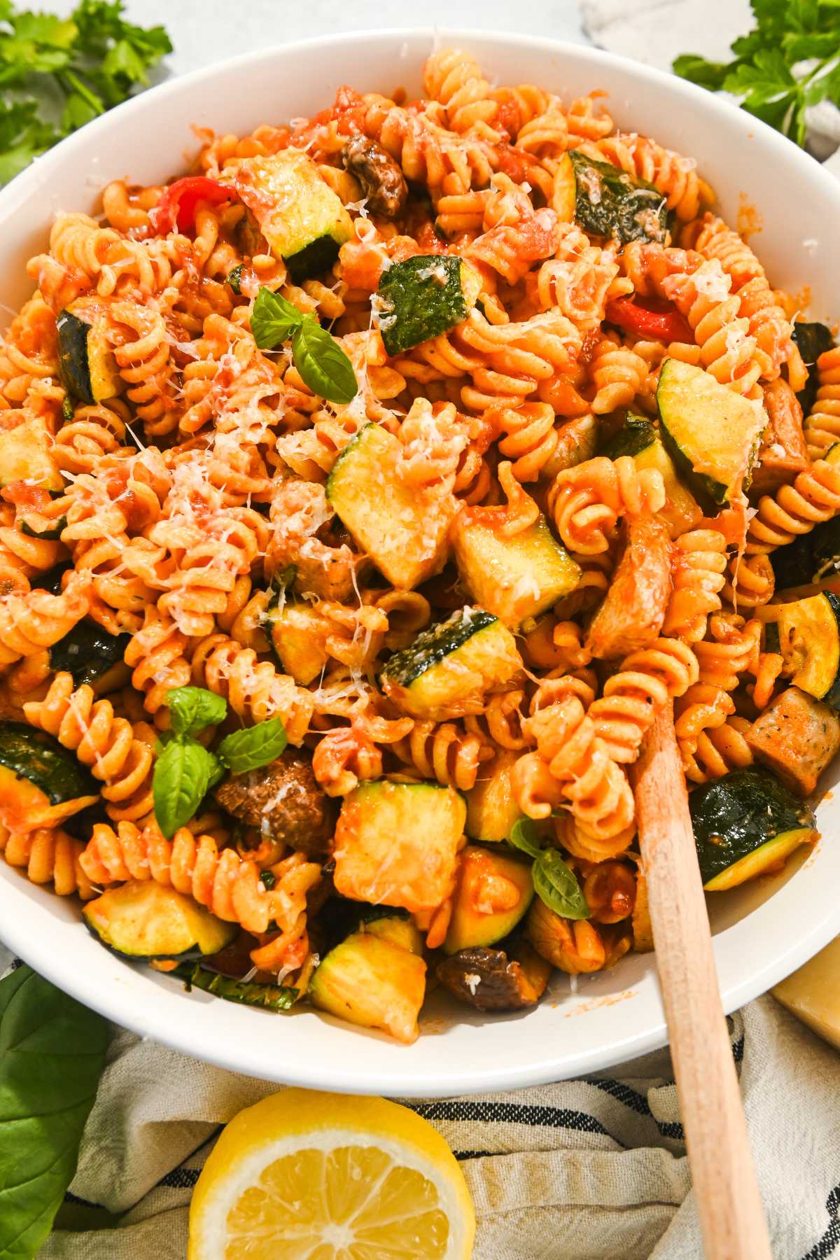 close up of chicken sausage pasta with chunks of grilled veggies served in a white bowl with a wooden spoon.