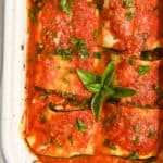 close up of six zucchini rolls in a white pan topped with sauce and basil.