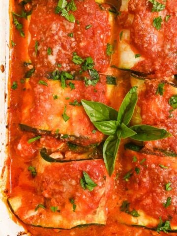 close up of six zucchini rolls in a white pan topped with sauce and basil.