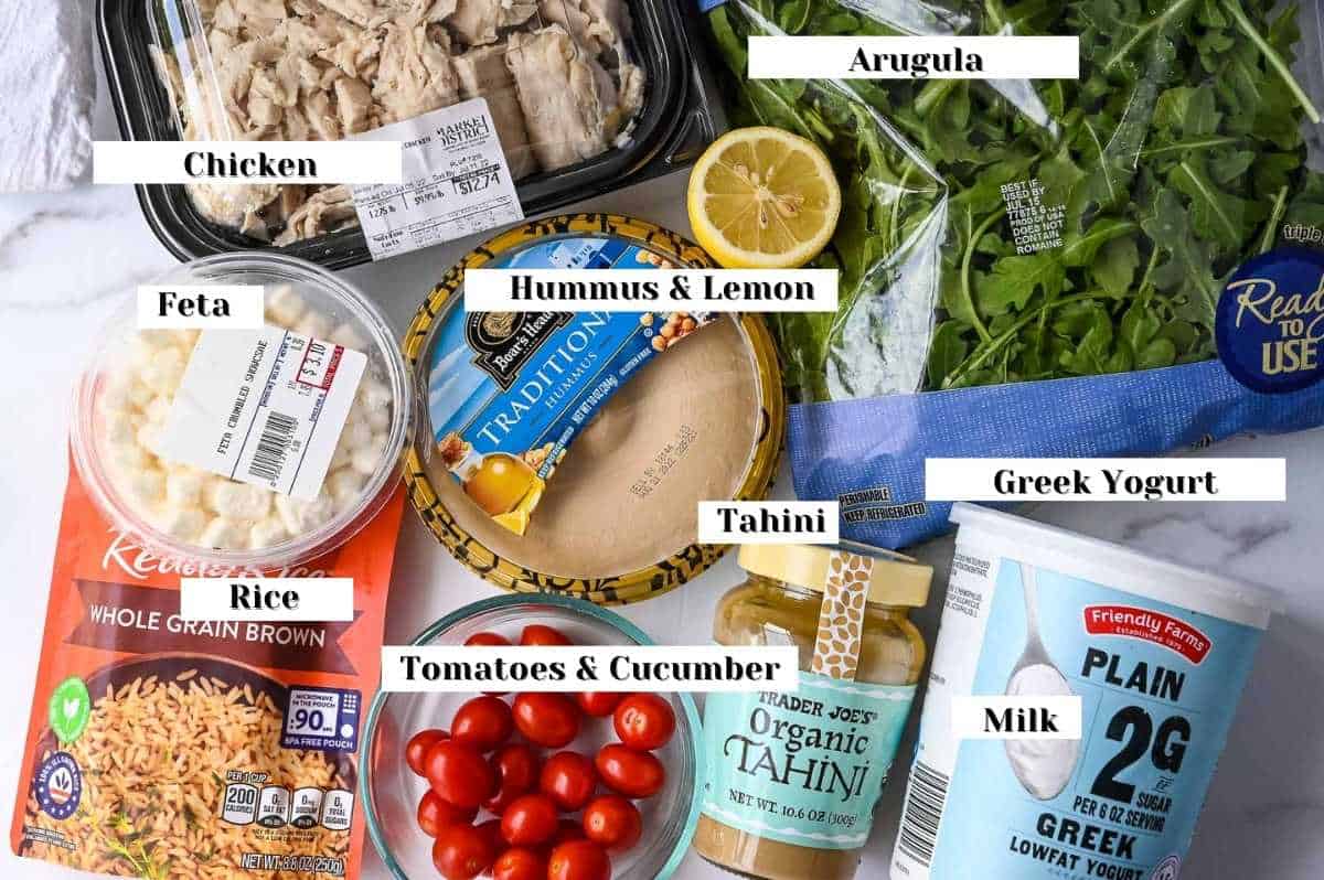 labeled ingredient shot of this recipe including chicken, arugula, Greek yogurt, tahini, lemon juice, tomatoes, humus, rice and feta.