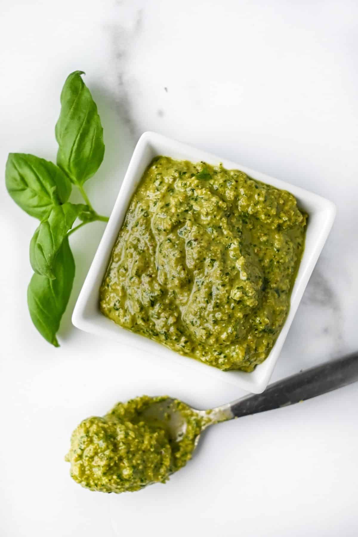 Full shot of dairy-free pesto in a white square dish next to a piece of fresh basil and a spoon, all on a white background.