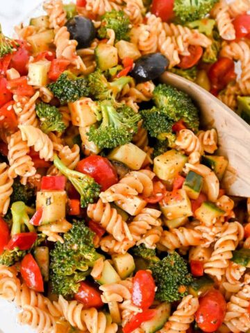 close up of gluten-free pasta salad with vegetables and a wooden spoon in a glass bowl.