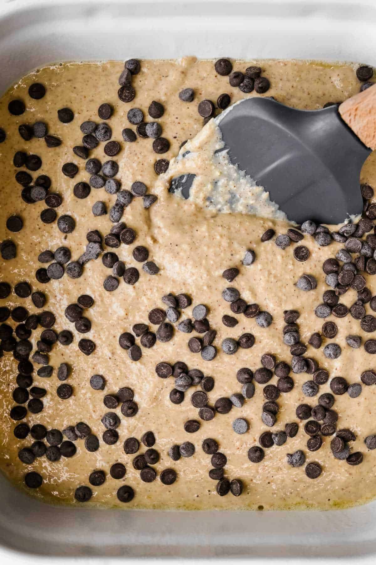 protein baked oatmeal in a white baking dish topped with chocolate chips with a spatula mixing the batter.