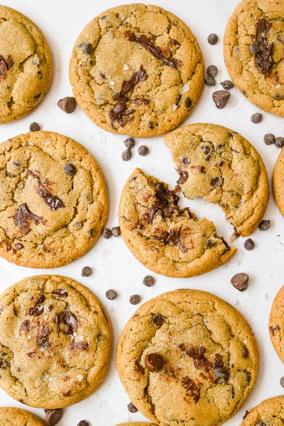 farther shot of batch of oat flour chocolate chip cookies with one broken in half on a white background with chocolate chips.
