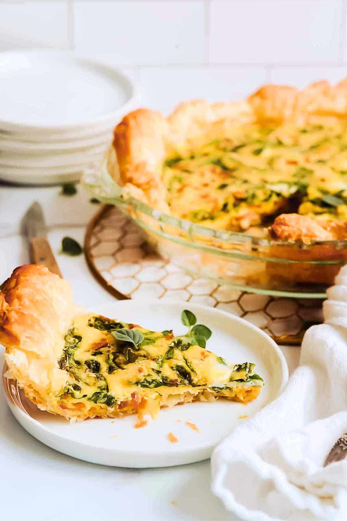 slice of puff pastry quiche on a white plate next to a whole pie plate of quiche.