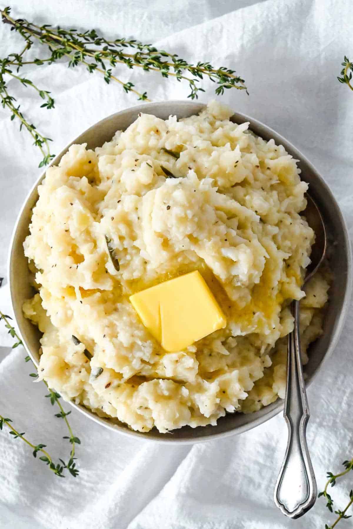 flat lay shot of mashed potatoes and cauliflower on a white background with a spoon in it and herbs around it.