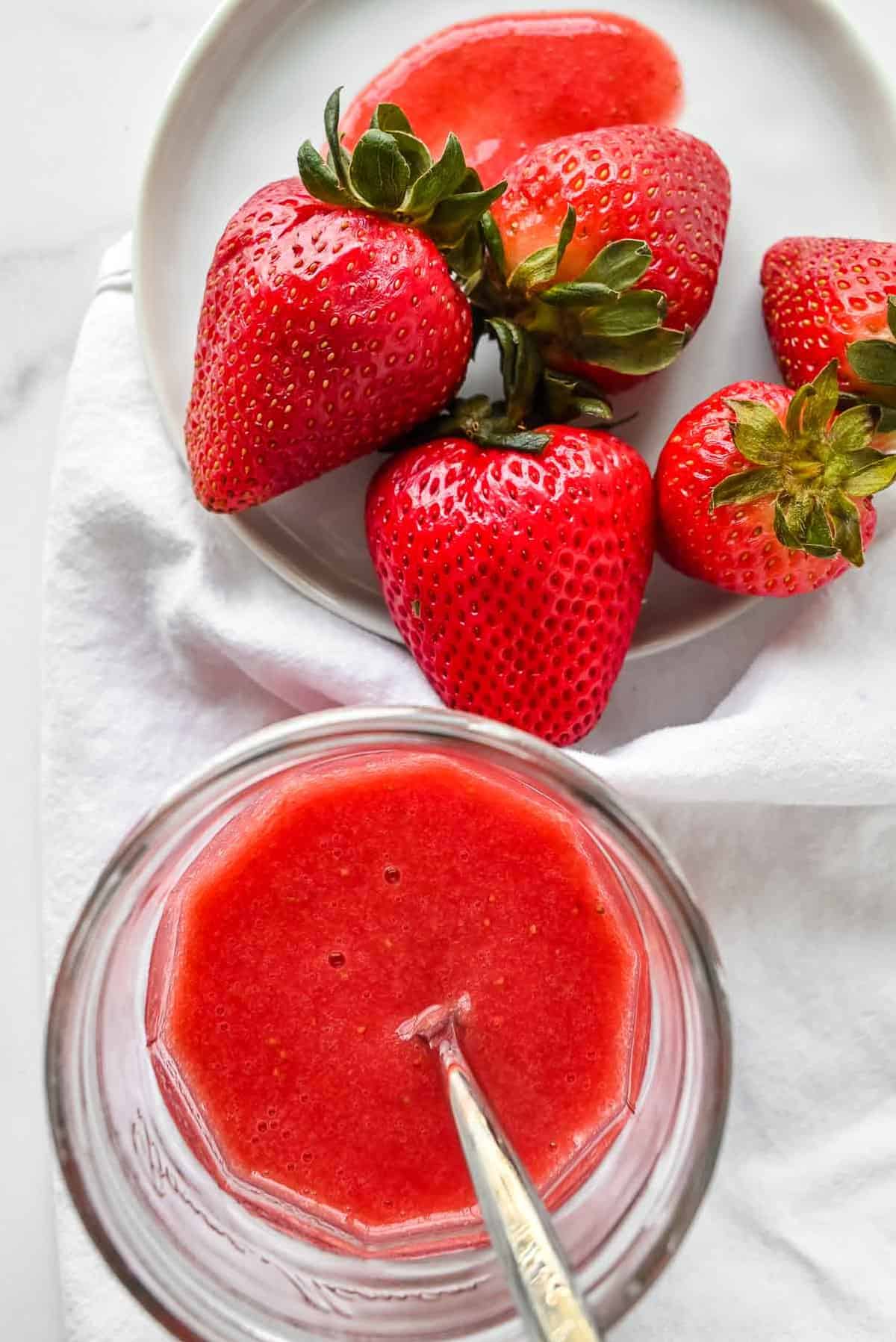 overhead shot of a bowl of strawberries next to strawberry puree in a glass jar with a spoon in it.