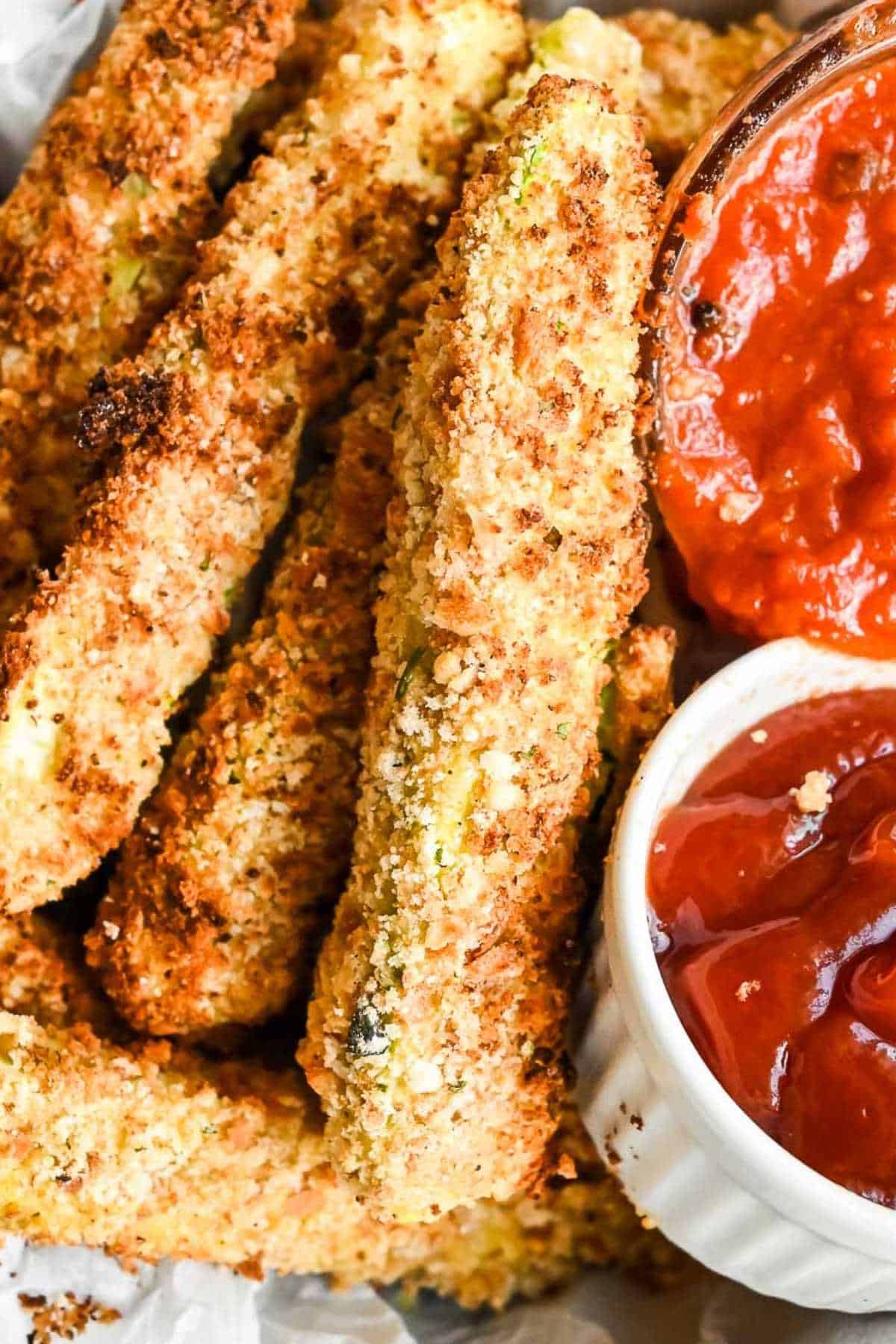air fryer zucchini sticks in a bowl with marinara and ketchup dipping sauces.