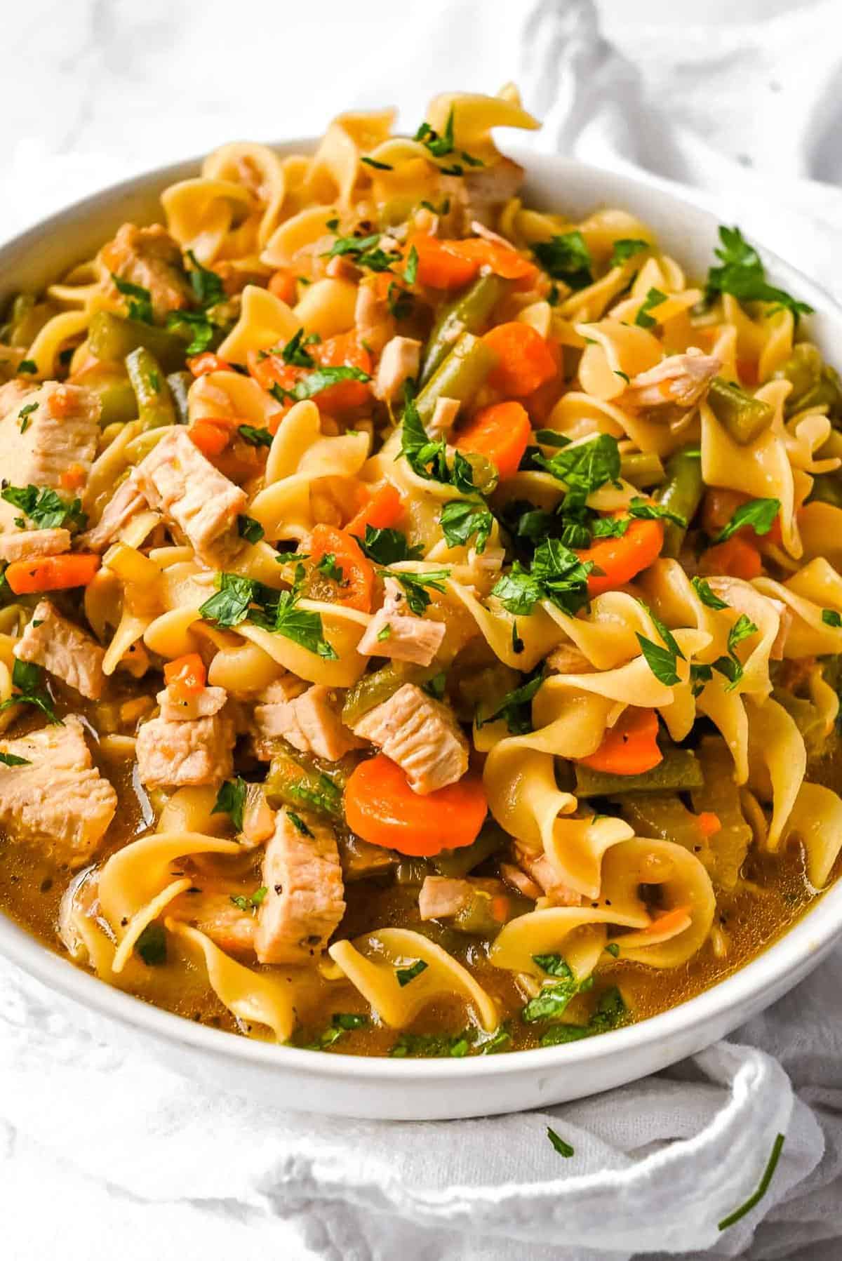 close up shot of turkey noodle soup in a white bowl on a white background.