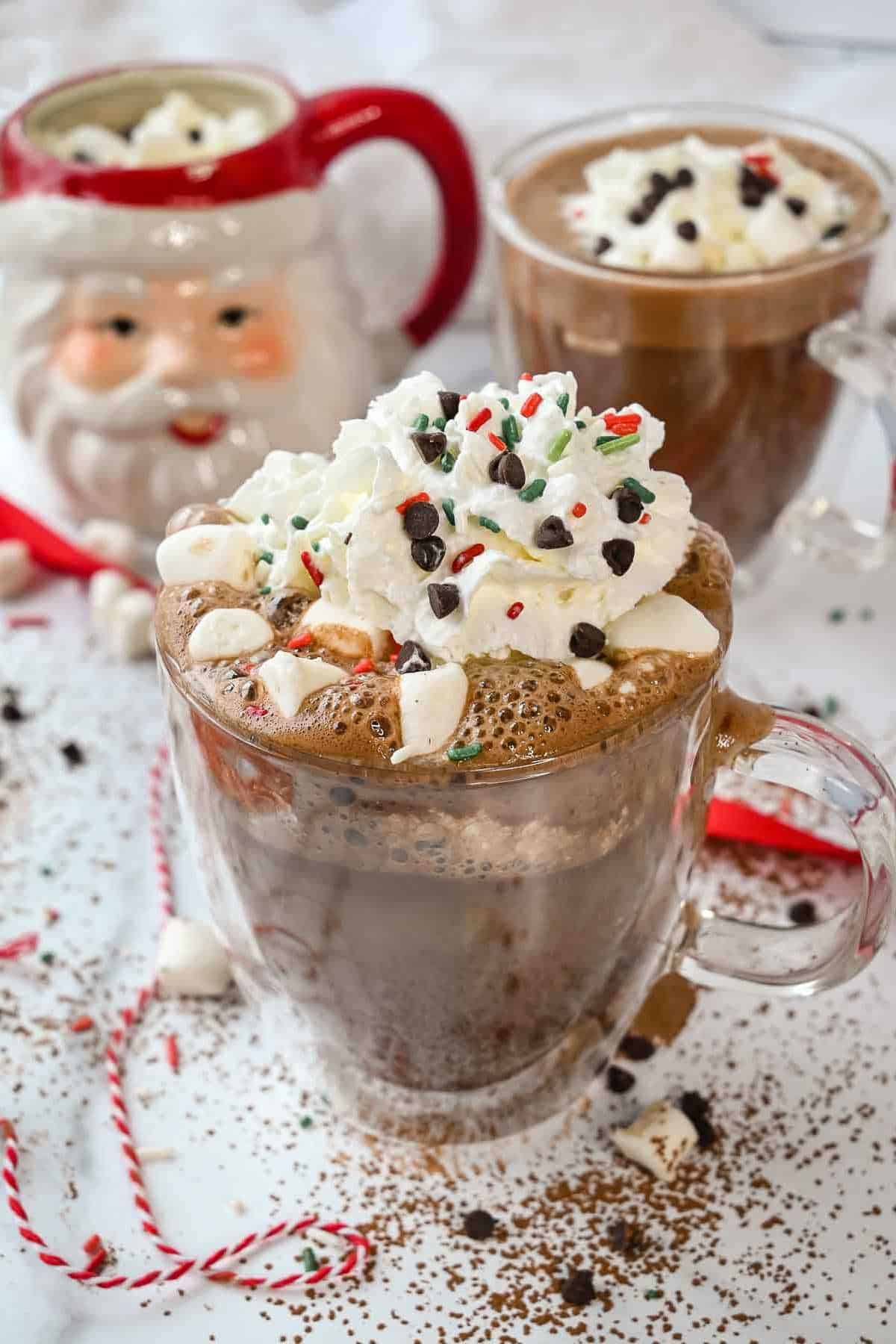 cup of oat milk hot chocolate with whipped cream, sprinkles, chocolate chips and marshmallows on a white background.