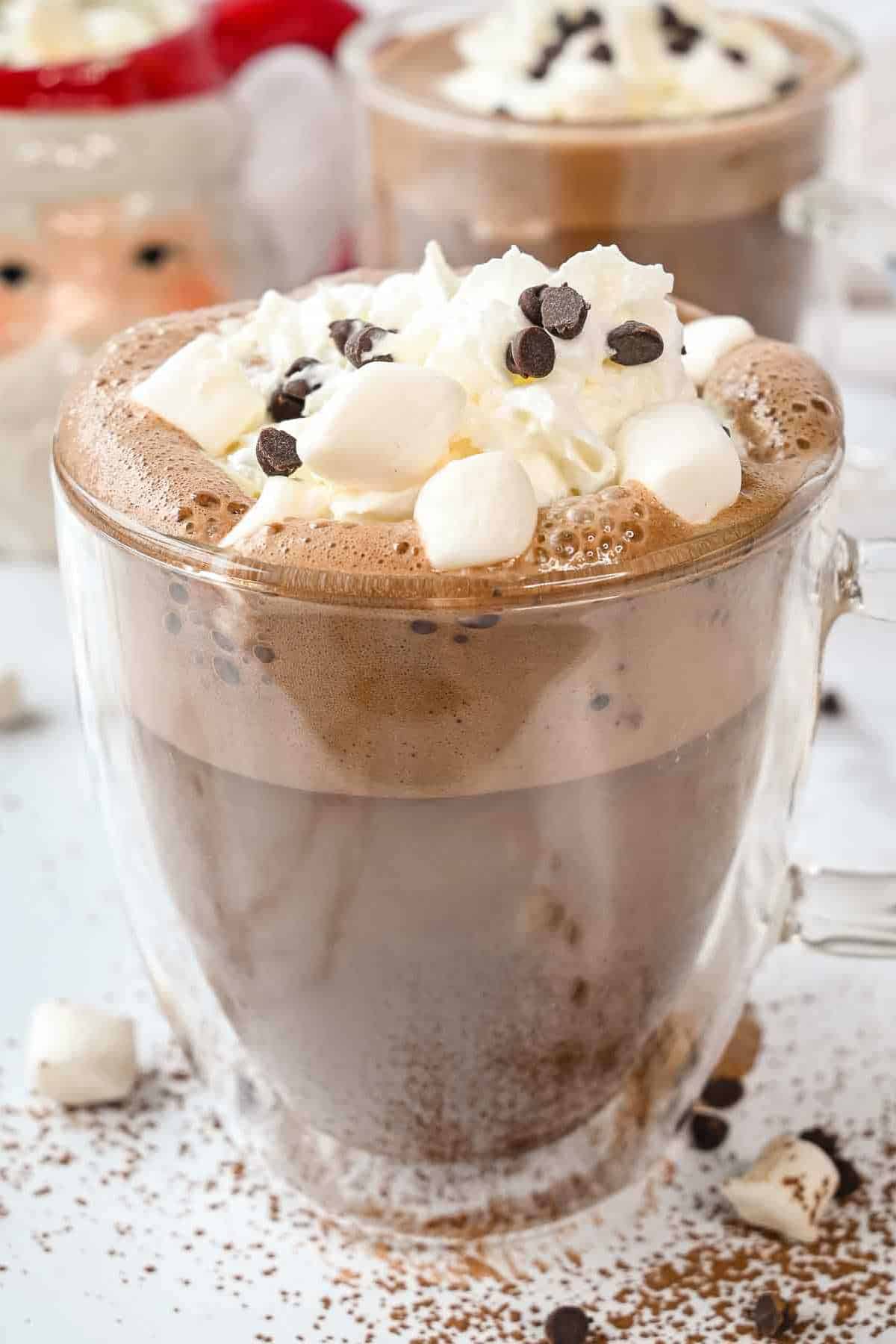 cup of oat milk hot chocolate with whipped cream, chocolate chips and marshmallows on a white background.