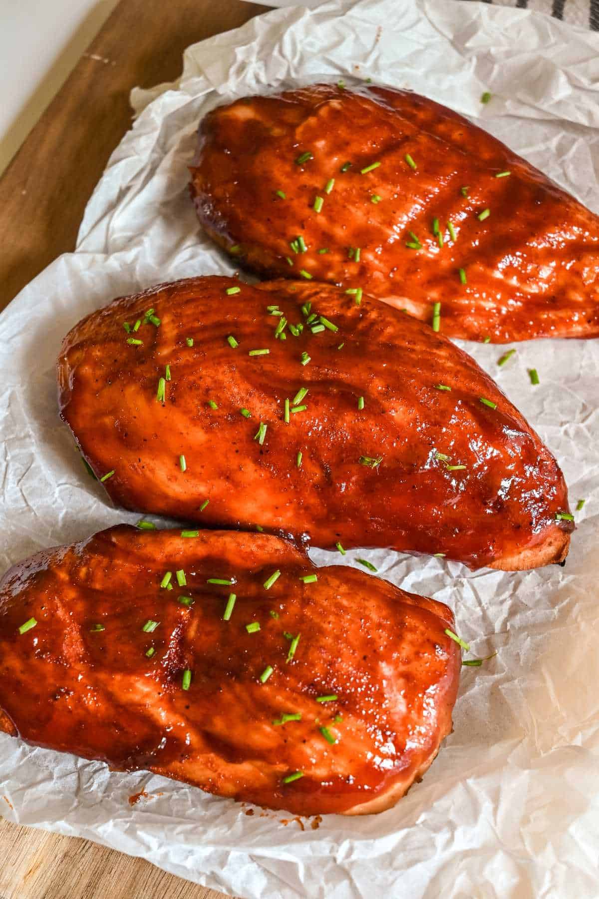 three bbq chicken breast on white parchment paper topped with chives.