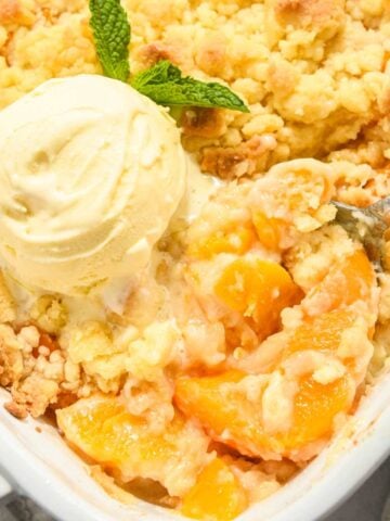 close up of peach cobbler in a white square dish with a scoop of ice cream on top and mint.