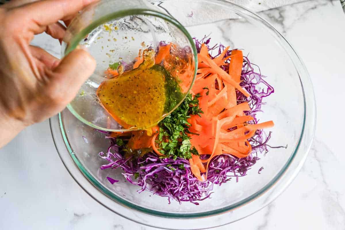 hand pouring dressing over red cabbage slaw in a glass bowl.