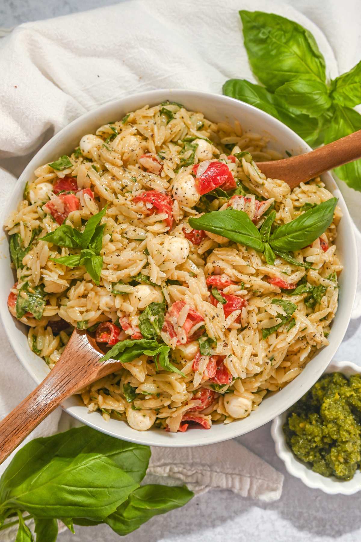 overhead view of pesto orzo salad in a white bowl with tomatoes, fresh mozzarella, and fresh basil with wooden utensils.