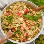 overhead view of pesto orzo salad with tomatoes, fresh mozzarella, and fresh basil in a white bowl with wooden utensils.