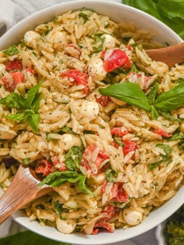 overhead view of pesto orzo salad with tomatoes, fresh mozzarella, and fresh basil in a white bowl with wooden utensils.