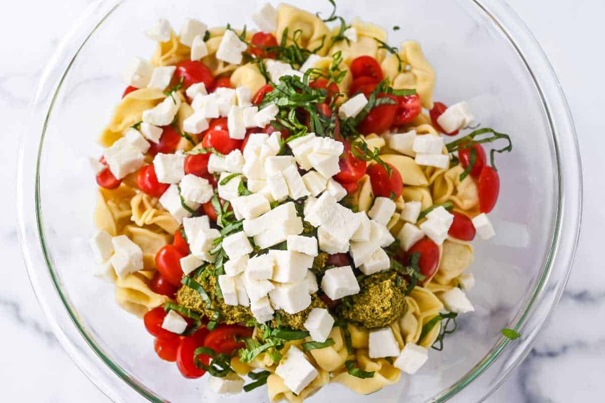 pesto tortellini salad ingredients in a glass bowl topped with basil and cheese.