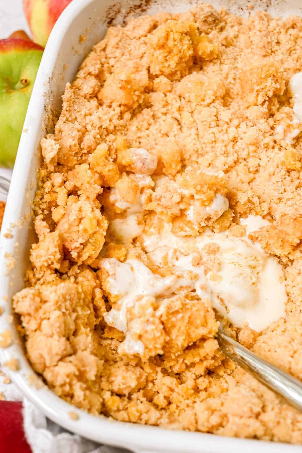 full view close up shot of apple cobbler with cake mix in a white dish topped with ice cream and a spoon in the dish.