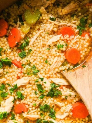 close up of chicken pastina soup with carrots and celery topped with parsley with a wooden spoon on top.