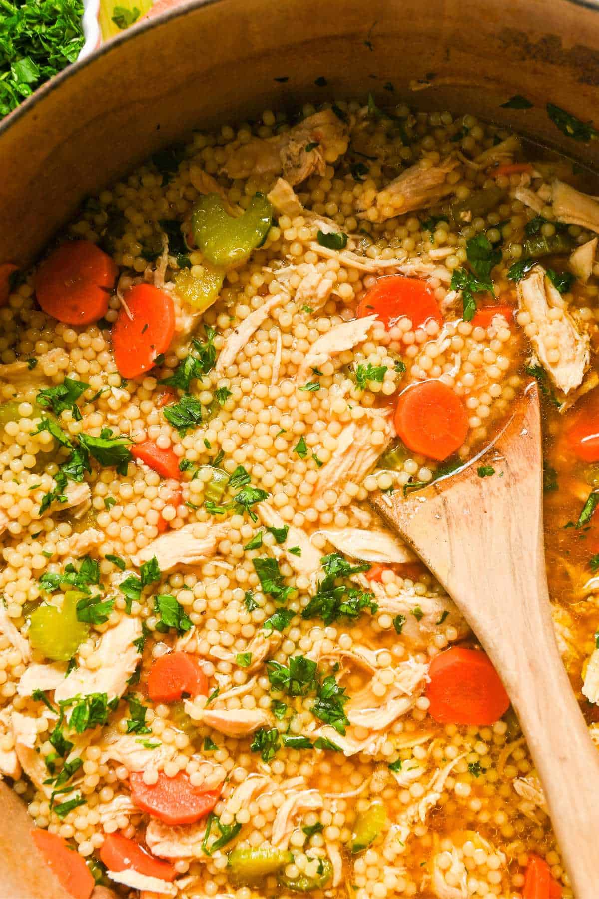 chicken pastina soup with carrots and celery topped with parsley with a wooden spoon on top.