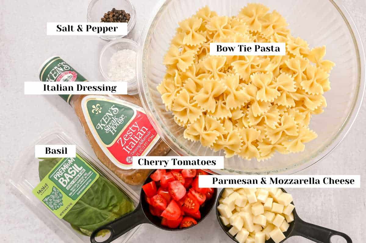 labeled ingredients for making caprese bow tie pasta salad on a gray background.