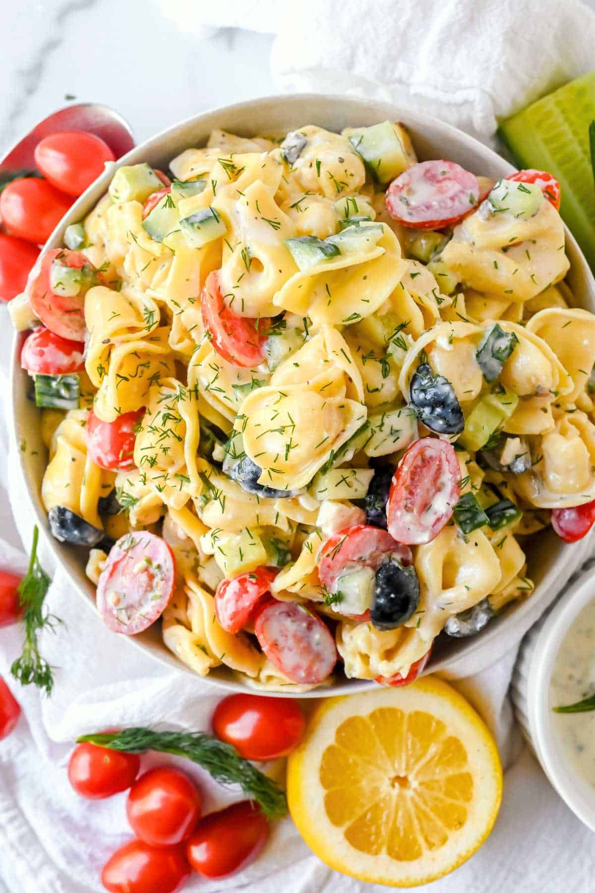 top down view of greek tortellini pasta salad with ingredients around it on a white background.
