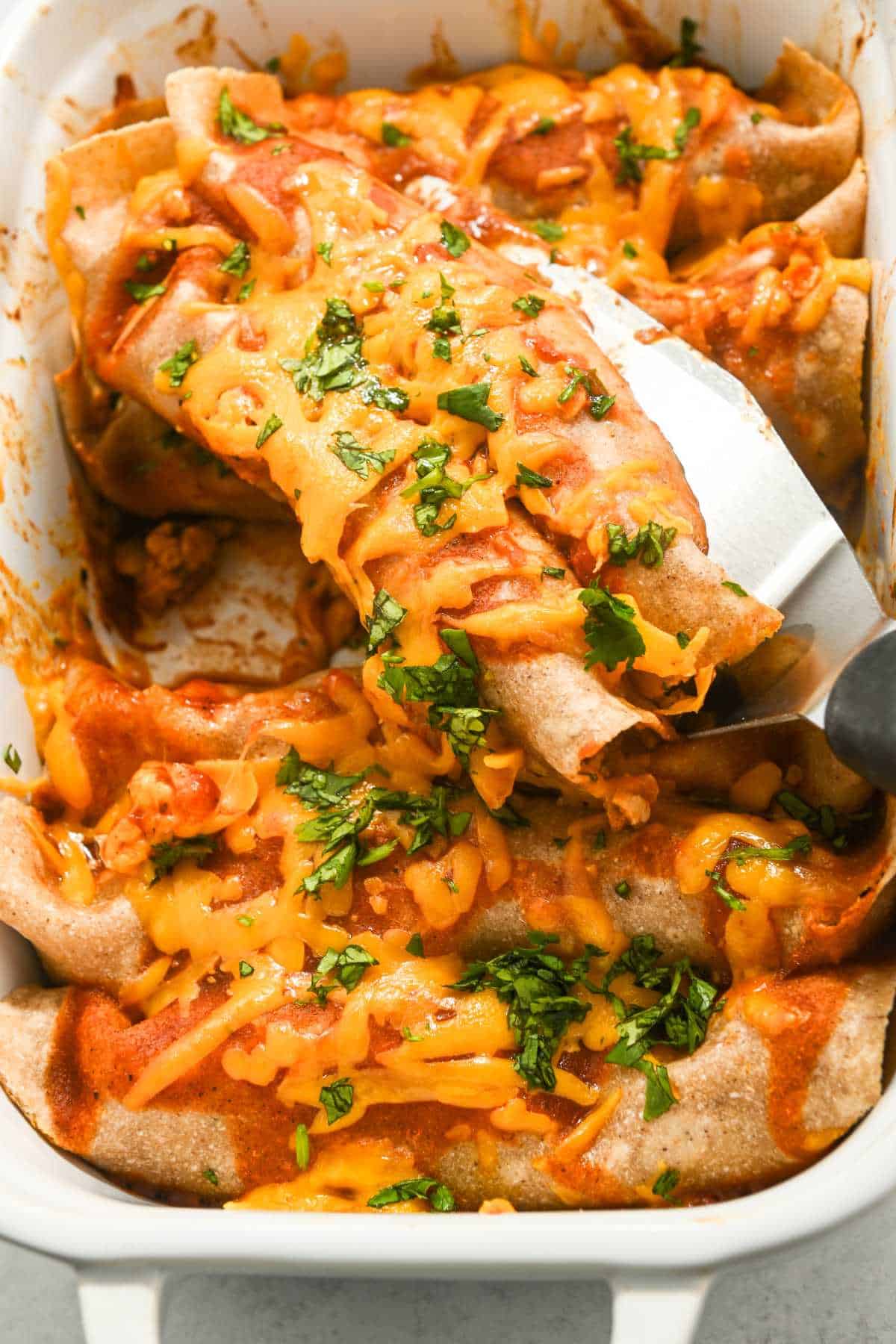 pan of ground chicken enchiladas with a spatula lifting two enchiladas out.