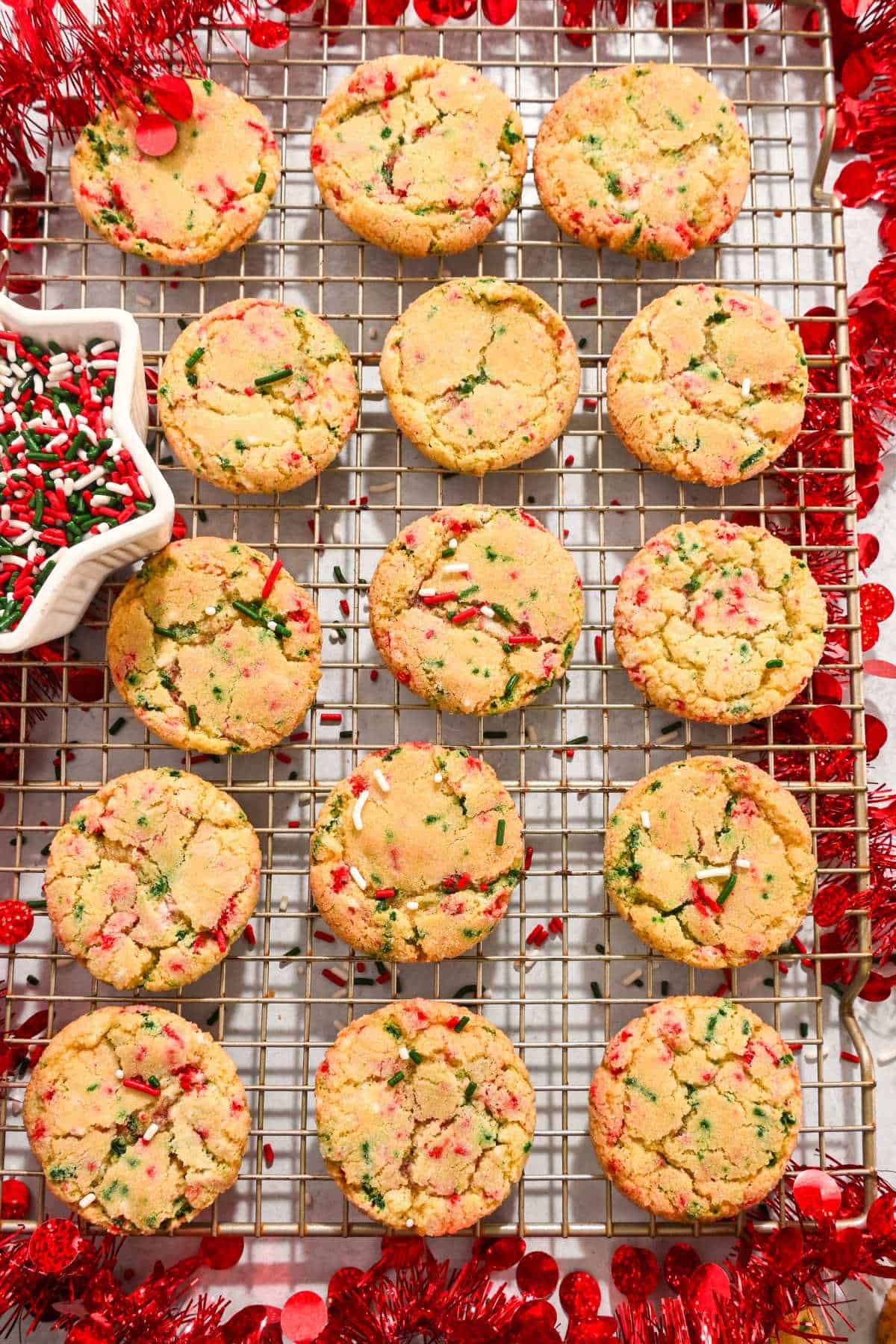 15 sugar cookies with christmas sprinkles on a cooling rack surrounded by tinsel.