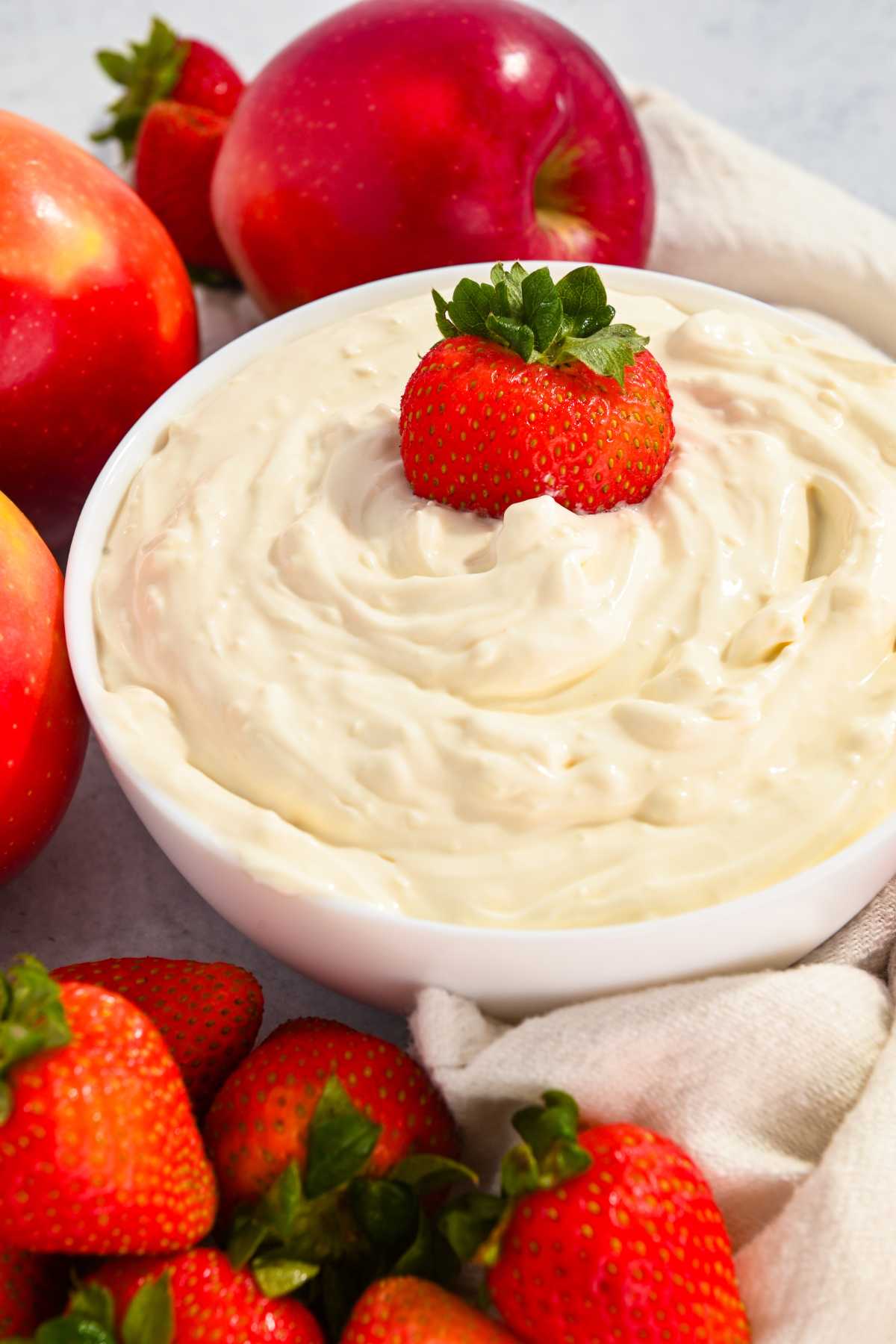a white bowl filled with healthy cheesecake dip with a strawberry on top and fruit around it.