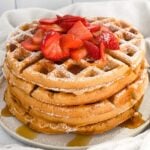 close up of stack of kodiak waffles topped with powdered sugar, syrup and strawberries on a white plate.