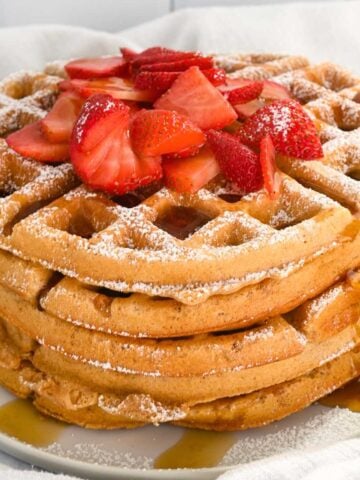 close up of stack of kodiak waffles topped with powdered sugar, syrup and strawberries on a white plate.