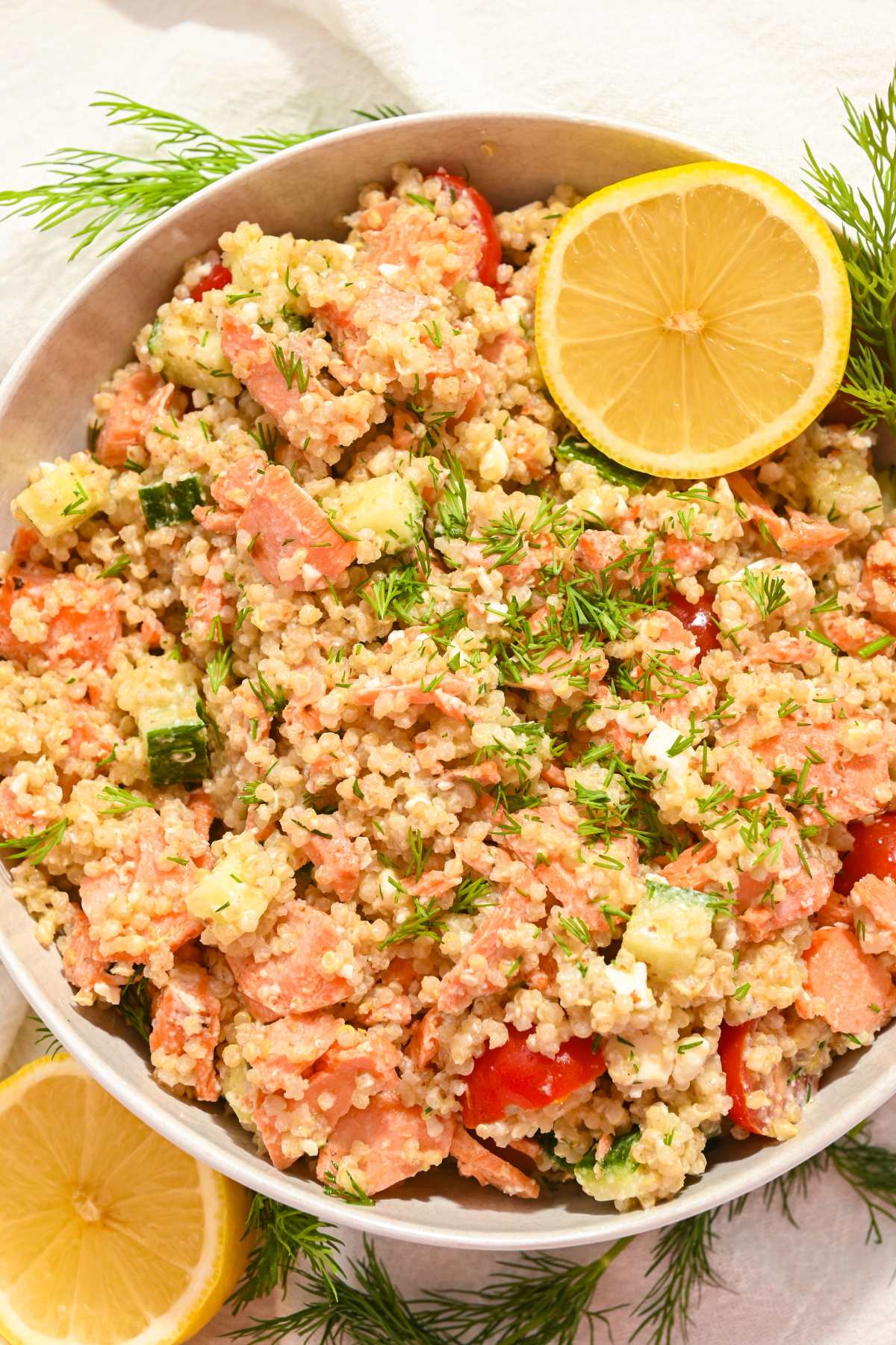 white bowl full of salmon quinoa salad with a lemon on top on a white background with lemons and dill underneath the bowl.