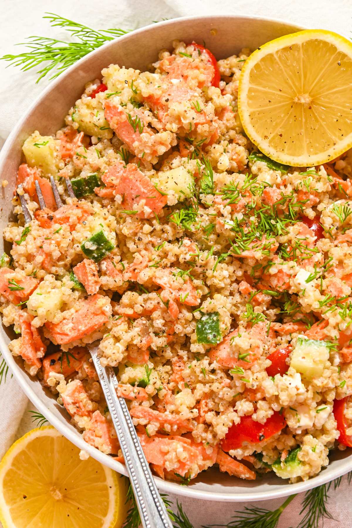 salmon quinoa salad with flaked salmon, diced cucumber, sliced cherry tomatoes, and fresh dill along with lemon slices in a white bowl with a fork.