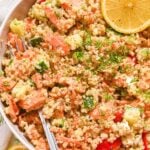 close up of salmon quinoa salad with flaked salmon, diced cucumber, sliced cherry tomatoes, and fresh dill along with lemon slices in a white bowl with a fork.