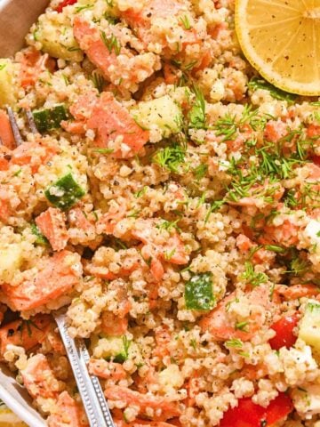 close up of salmon quinoa salad with flaked salmon, diced cucumber, sliced cherry tomatoes, and fresh dill along with lemon slices in a white bowl with a fork.