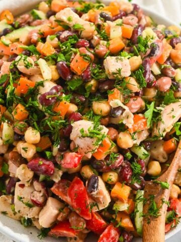 close up of a bowl of dense bean salad with chicken in a white bowl with a wooden spoon.