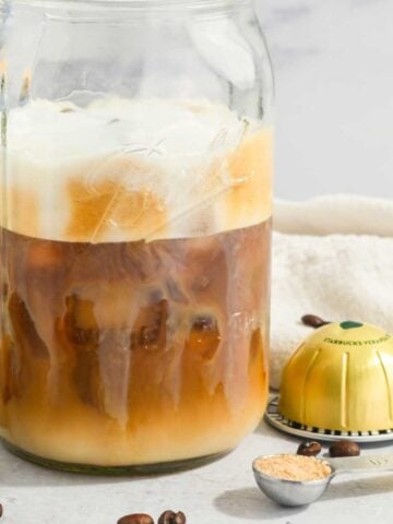 close up of Nespresso coffee in a mason jar with foam on top and with capsules, brown sugar, and coffee beans around it.