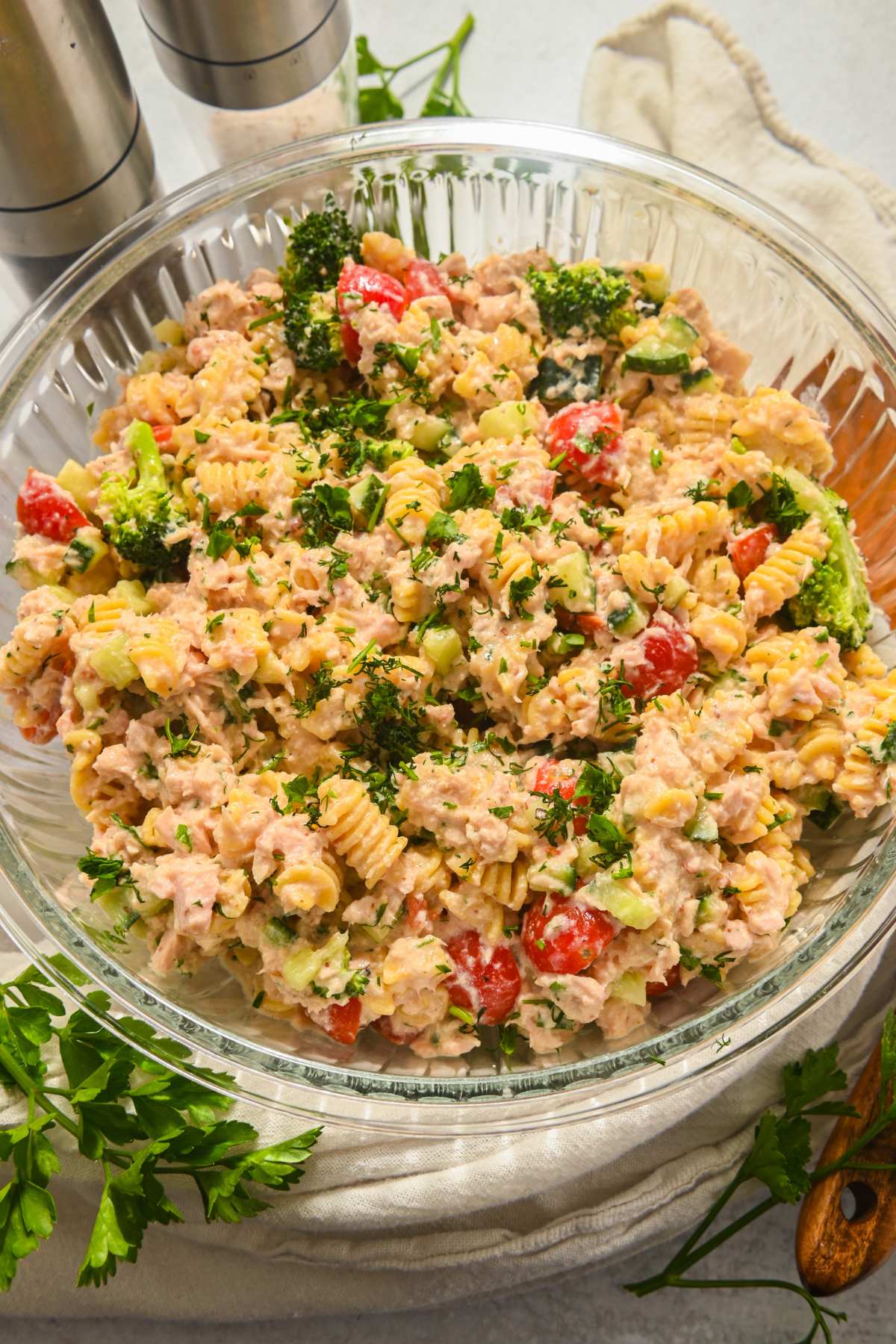 full view of high protein tuna pasta salad in a glass bowl with fresh herbs and salt and pepper around it.