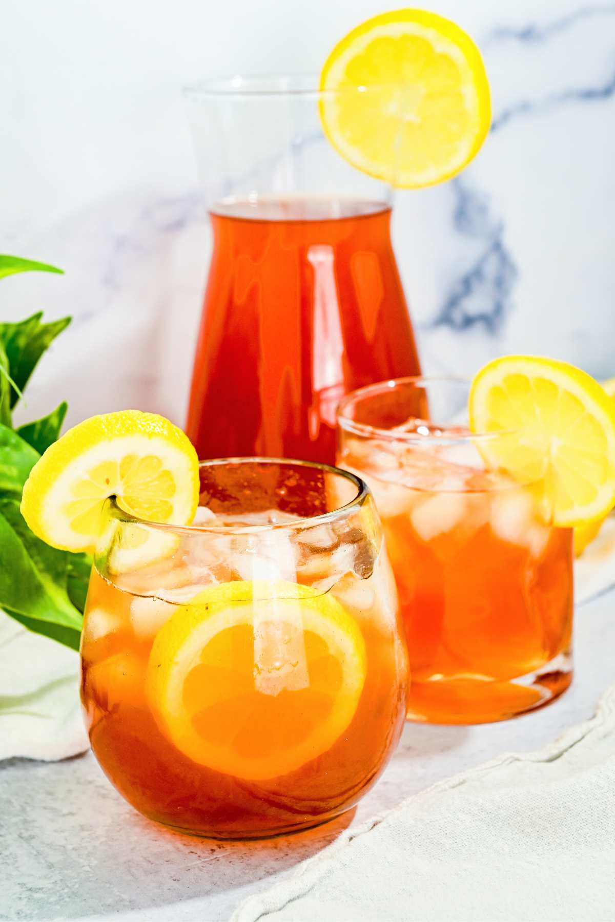 two glass of iced tea lemonade with lemons on the side in front of a pitcher of iced tea in the back.