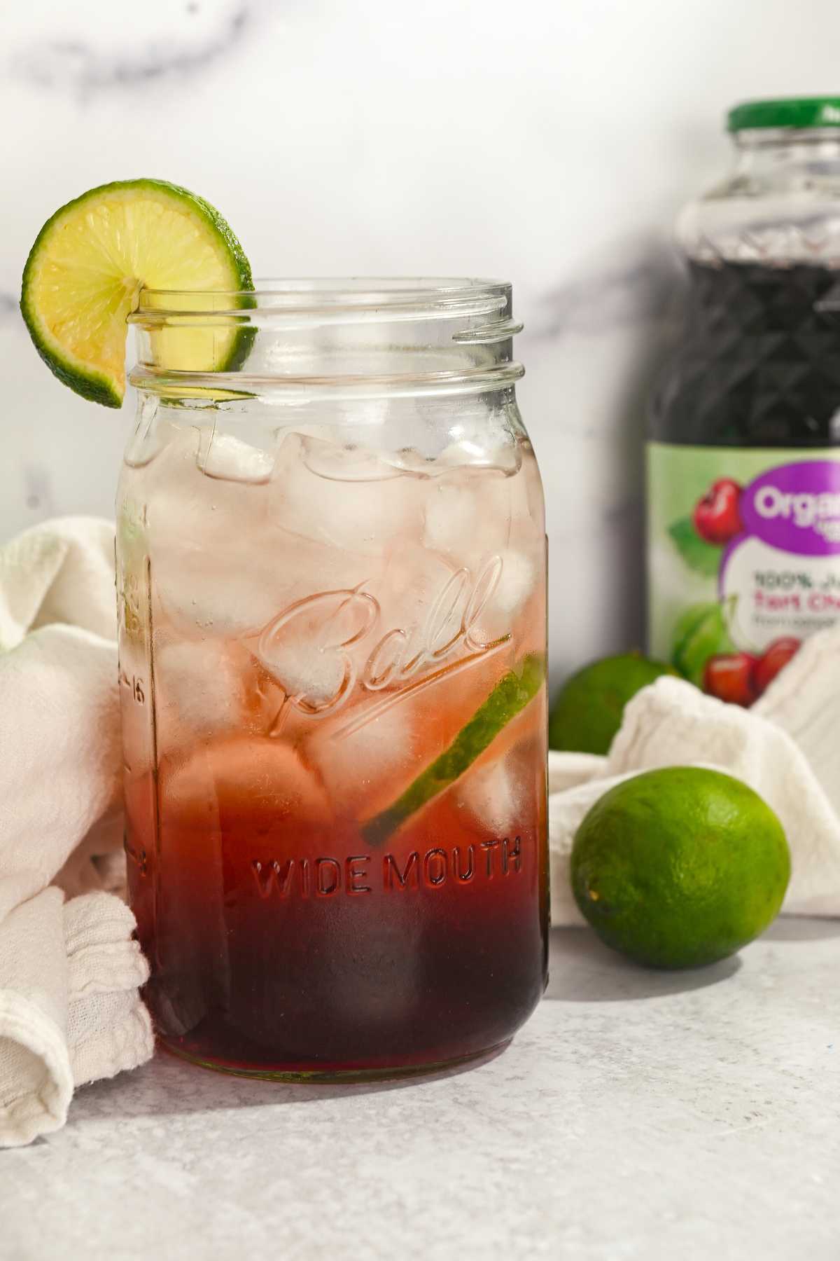 Iced tart cherry mocktail in a glass mason jar with a lime and jar of juice next to it.