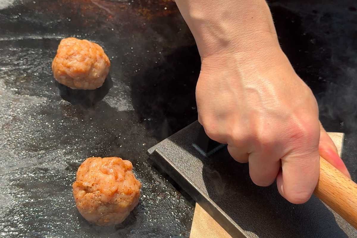hand pressing down a turkey burger ball on a hot Blackstone griddle using a burger press and parchment paper.