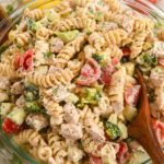 close up of creamy protein pasta salad with chicken, rotini, tomatoes, cucumbers, and broccoli in a glass bowl with a wooden spoon.