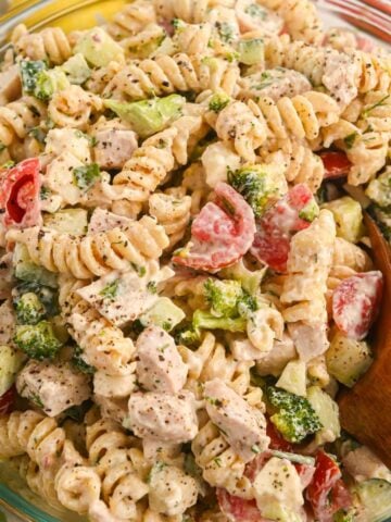 close up of creamy protein pasta salad with chicken, rotini, tomatoes, cucumbers, and broccoli in a glass bowl with a wooden spoon.