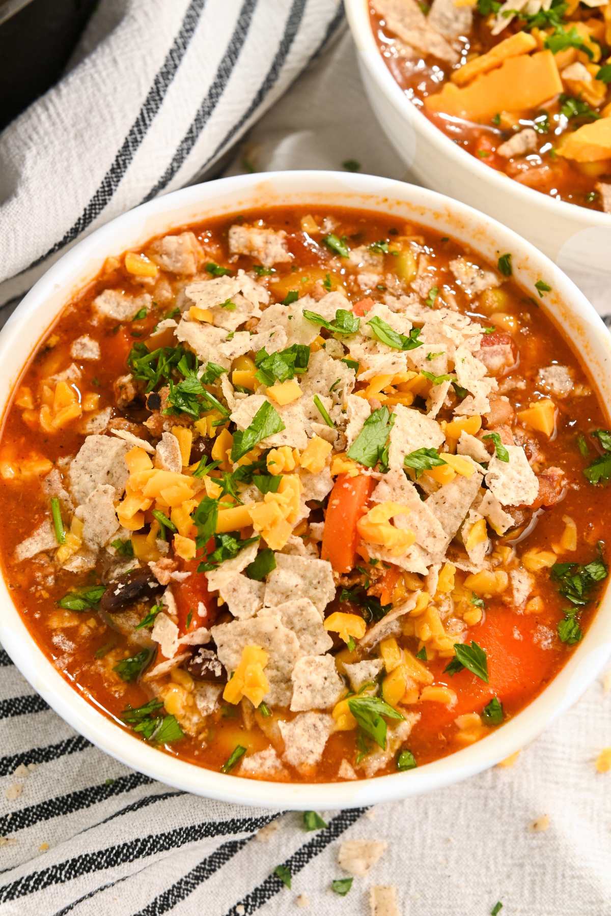 high protein chili in a white bowl on a blue and white striped towel next to another bowl of chili and covered with cheese, cilantro, and tortilla chip toppings.