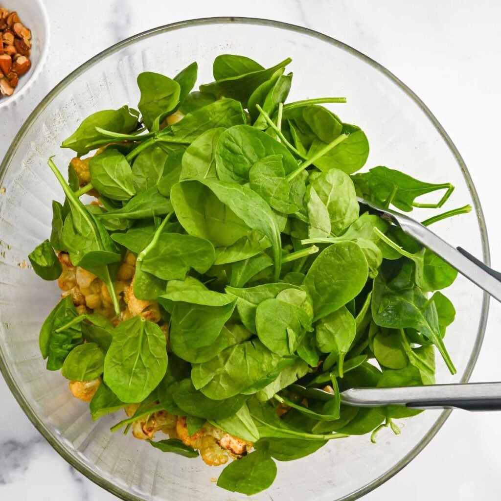 spinach on top of cauliflower salad in a glass bowl with tongs.