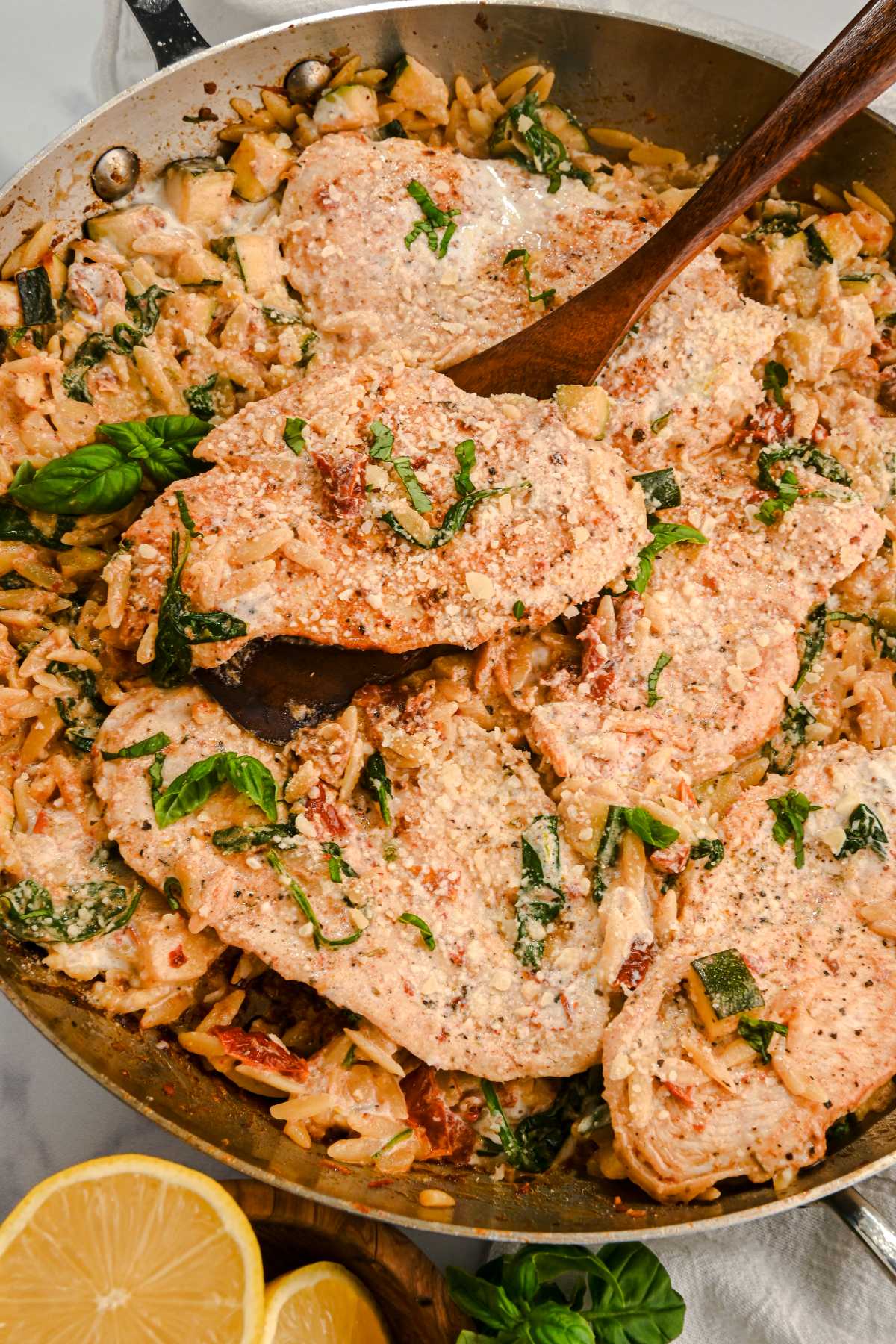 creamy orzo with chicken in a skillet with a wooden spatula, topped with basil and with lemon.